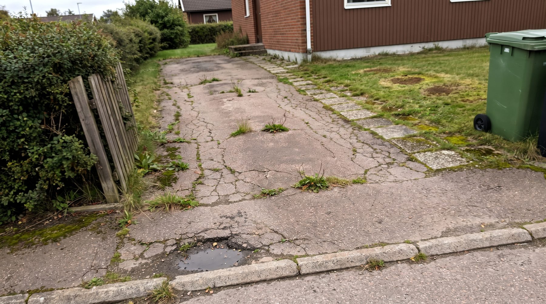 Sprucken asfaltsinfart med ogräs, pölar och en lutande staketstolpe.